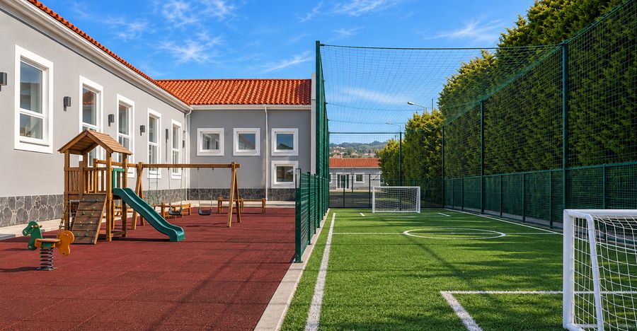 Parque infantil sobre piso vermelho ao lado de um campo de futebol de relva sintética vedado
