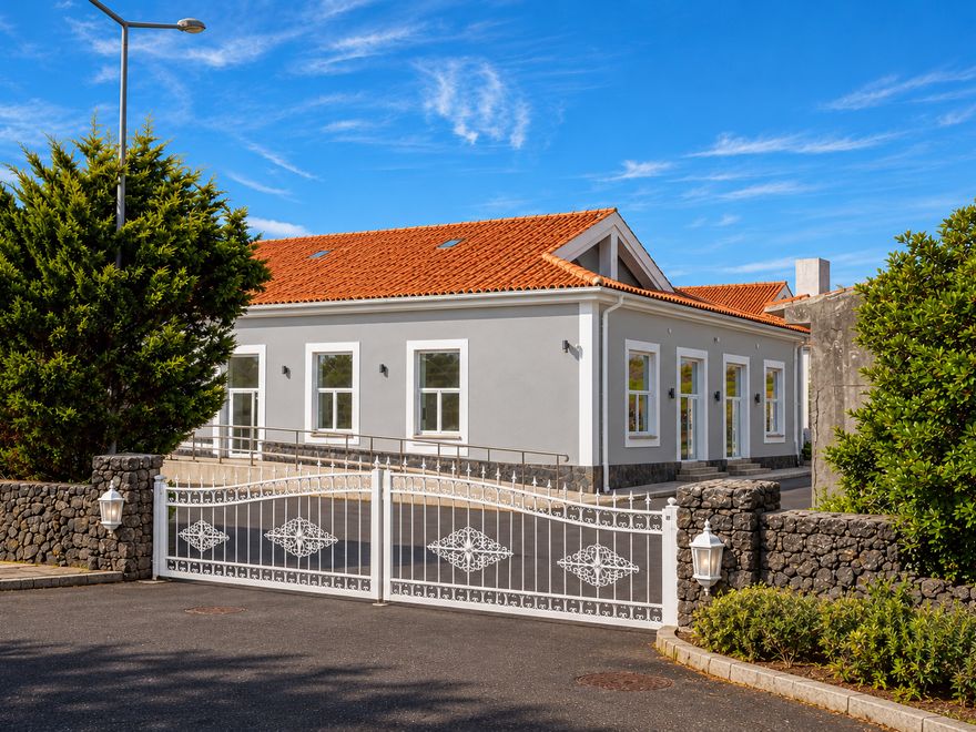 Portão branco ornamental entre muros de pedra vulcânica, com a fachada cinzenta ao fundo
